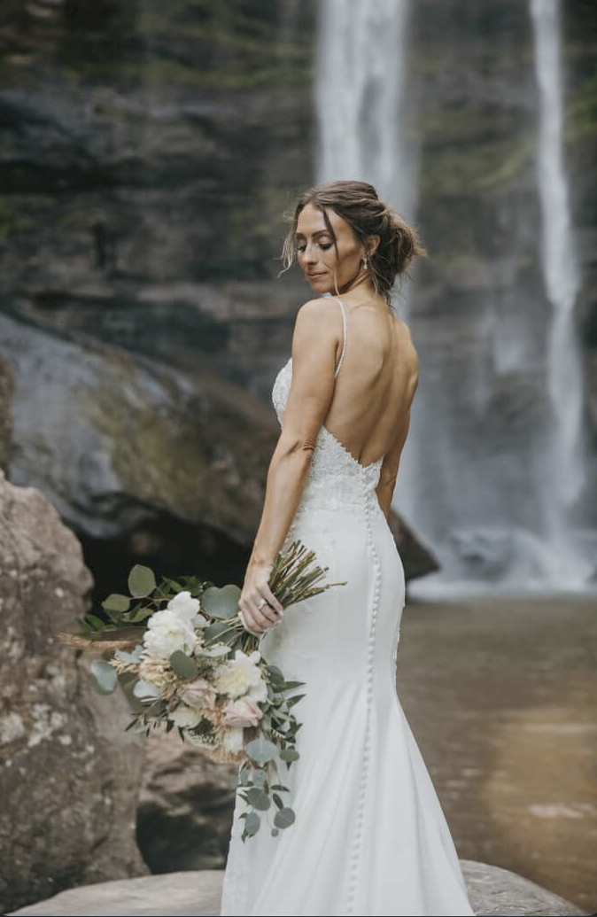 Sister Sunless bride in a backless wedding gown beside a waterfall